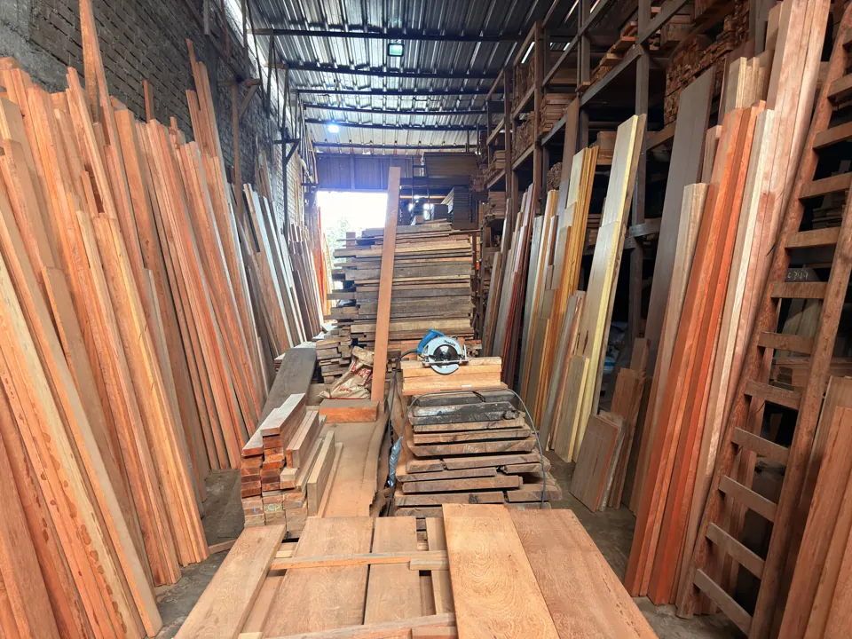 Pasillo central del almacén: tablones de pie a los lados, rumas de tablas, una escalera de madera y la luz que entra por el techo de calamina.